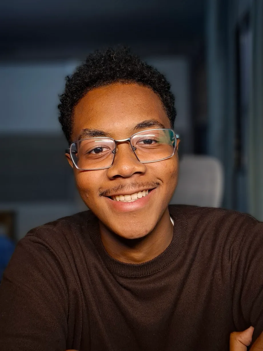 Retrato de Matheus Lucindo sorrindo, de óculos e blusa marrom